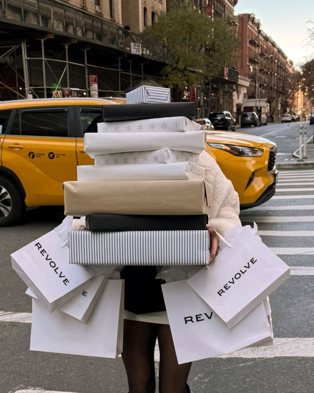 A person crossing a New York City street while carrying a massive stack of gift boxes and white Revolve shopping bags, with a yellow cab behind them.
