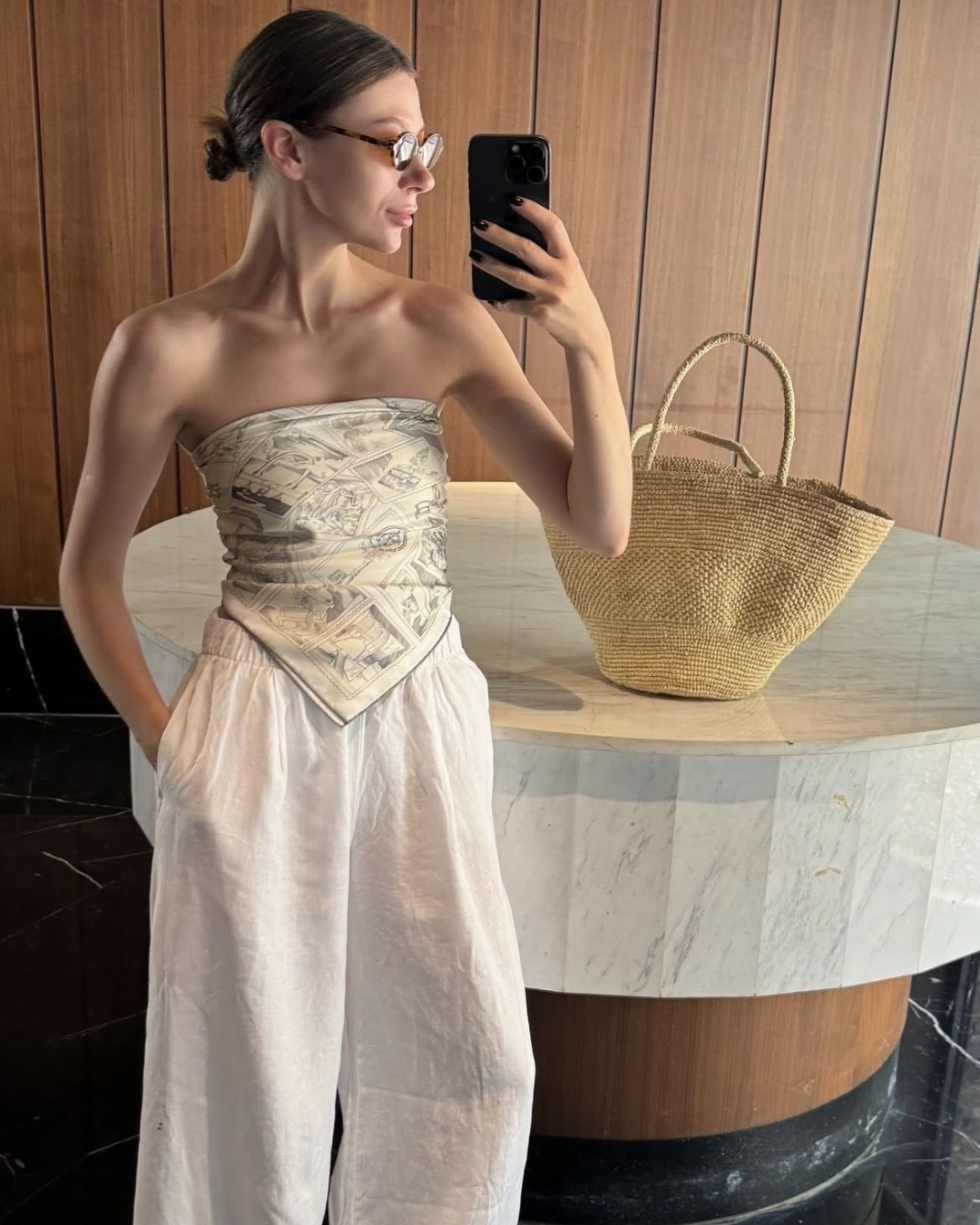 Effortless Mediterranean vacation style featuring a strapless geometric scarf top, loose white linen trousers, and a woven raffia basket tote bag