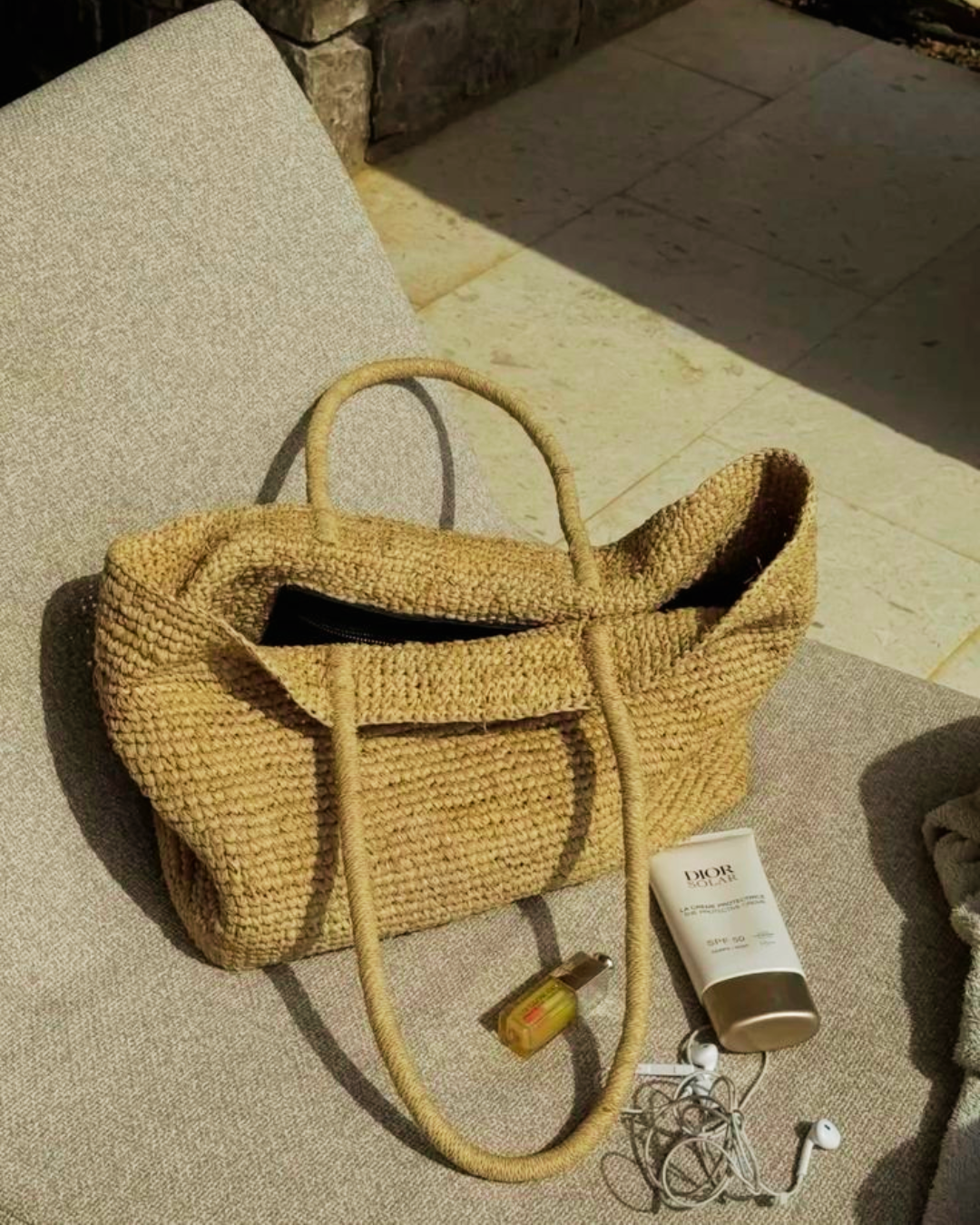 A structured woven jute straw tote bag placed on a lounge chair next to a tube of sunscreen and earphones under the sun.