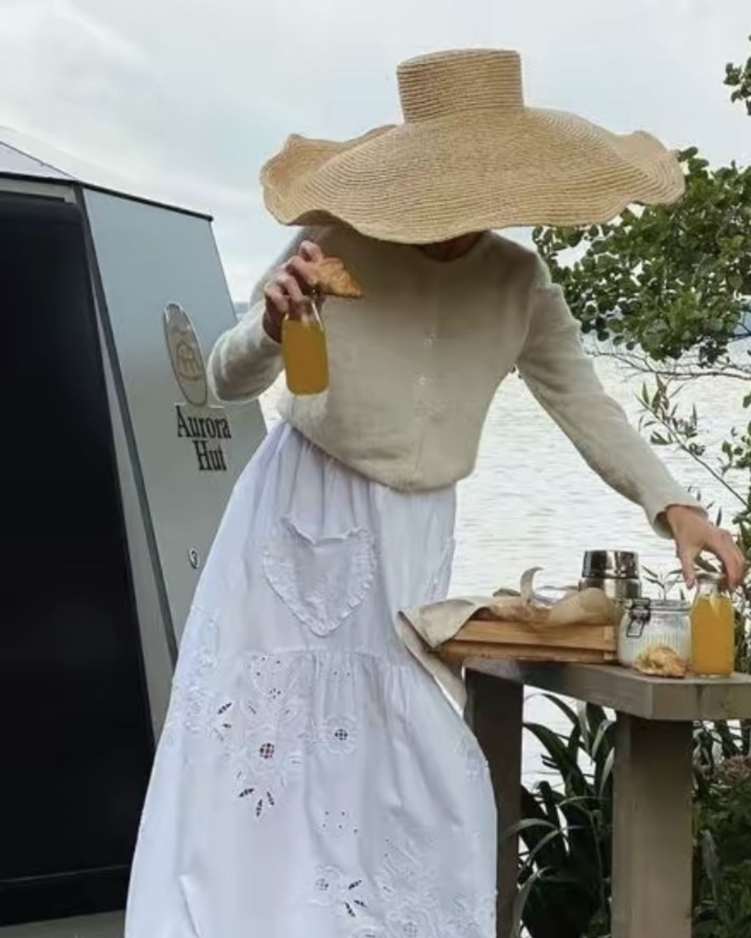 A woman in an oversized wide-brimmed straw hat and a white embroidered maxi skirt setting a picnic table by the water.