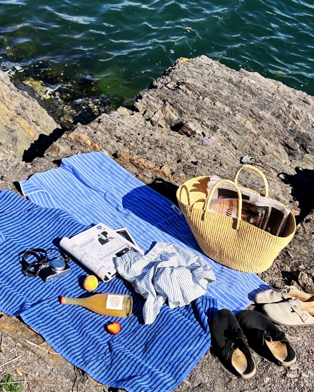 Minimalist summer picnic setup on coastal rocks featuring blue striped towels, a woven raffia basket bag with newspapers, summer slides, and a vintage camera