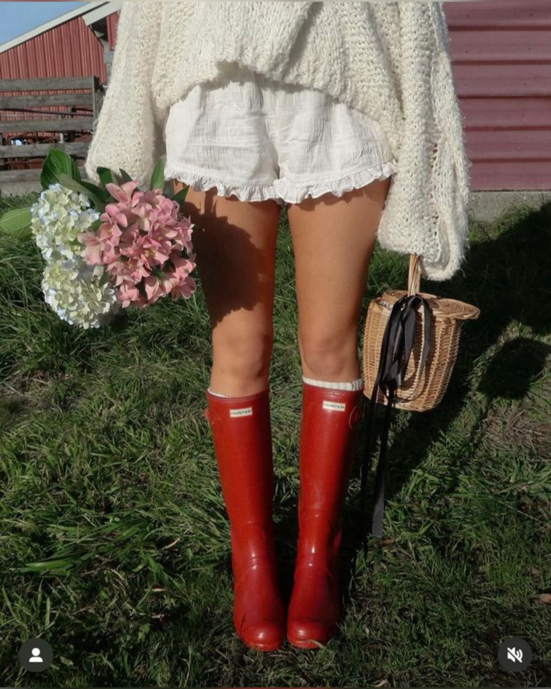 A person wearing bright red Hunter rubber rain boots and white ruffled shorts, holding flowers and a woven basket on a green grass field.