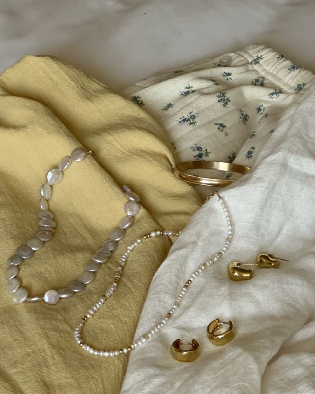Flat lay of summer fashion items including a yellow linen top, a baroque pearl necklace, a beaded pearl necklace, and chunky gold hoop earrings on a bed.