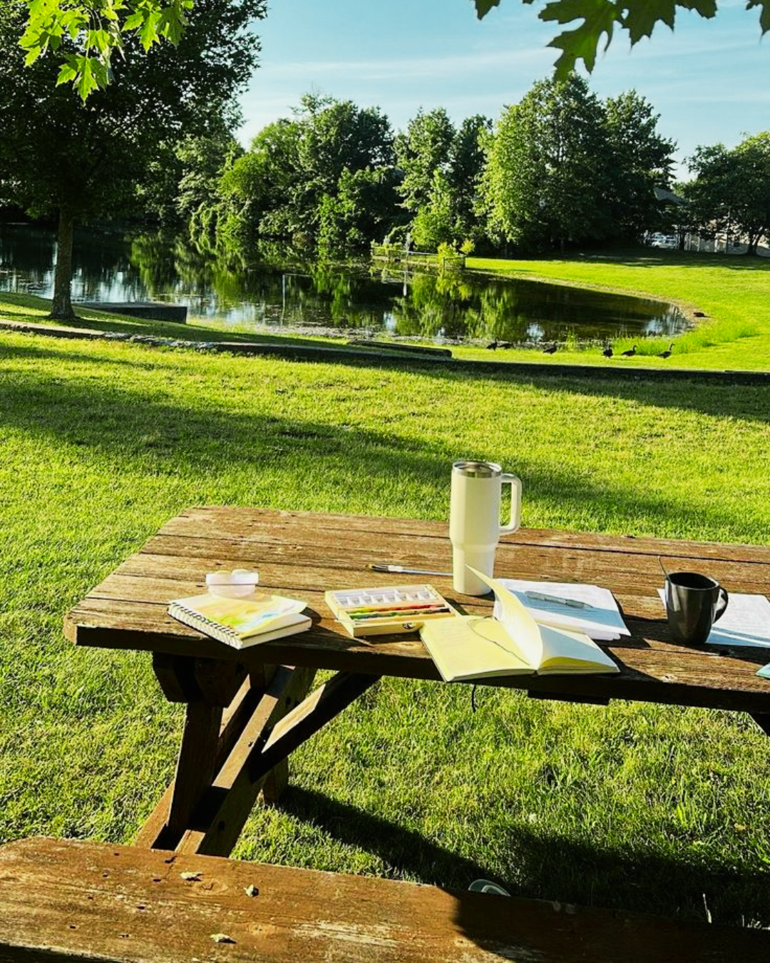 A wooden picnic table on a green lawn with a scenic lake and lush trees in the background, featuring notebooks, a watercolor palette, and a tumbler.