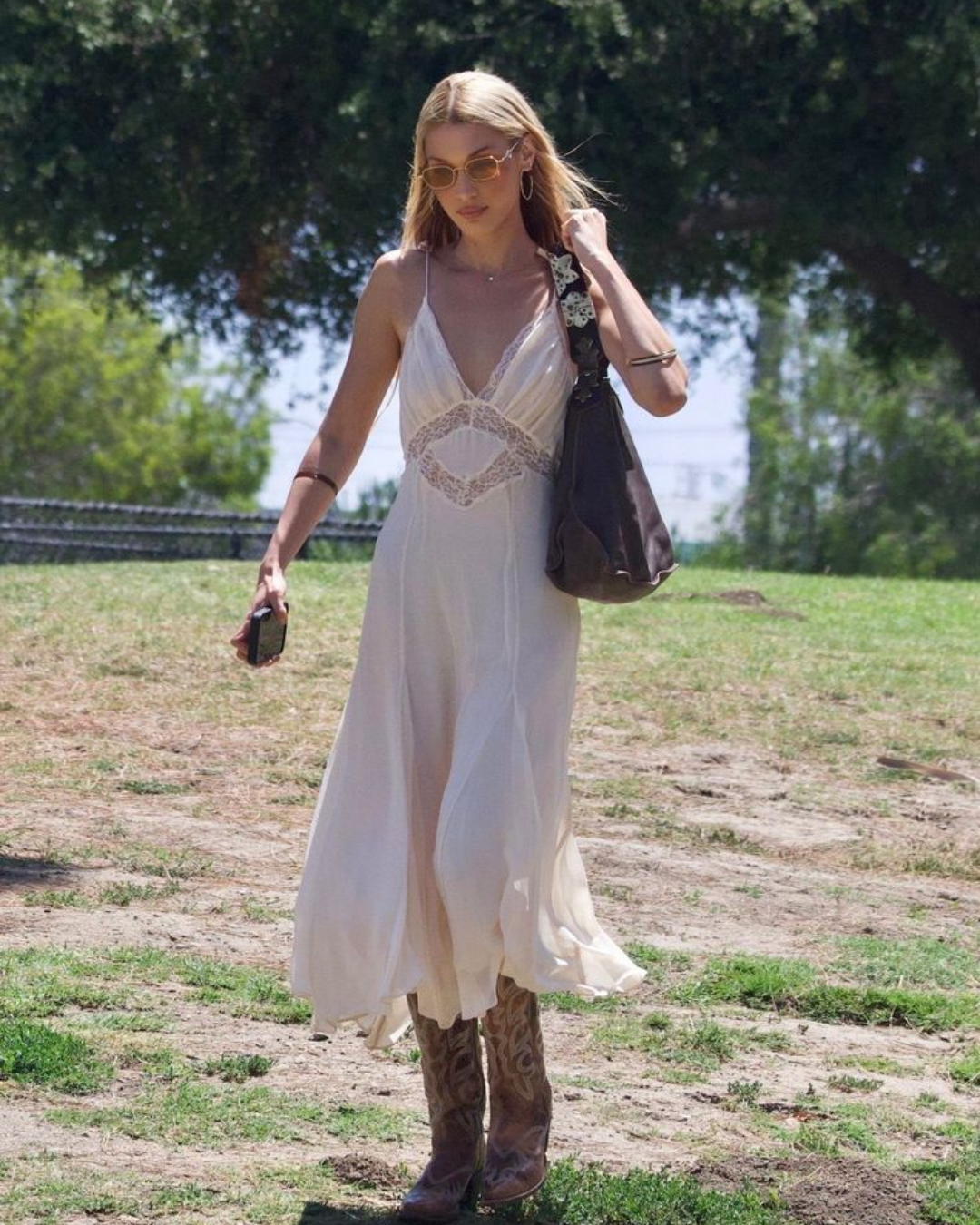 A blonde woman wearing a flowy cream lace maxi slip dress, sunglasses, and embroidered leather boots walking on an open grassy field.